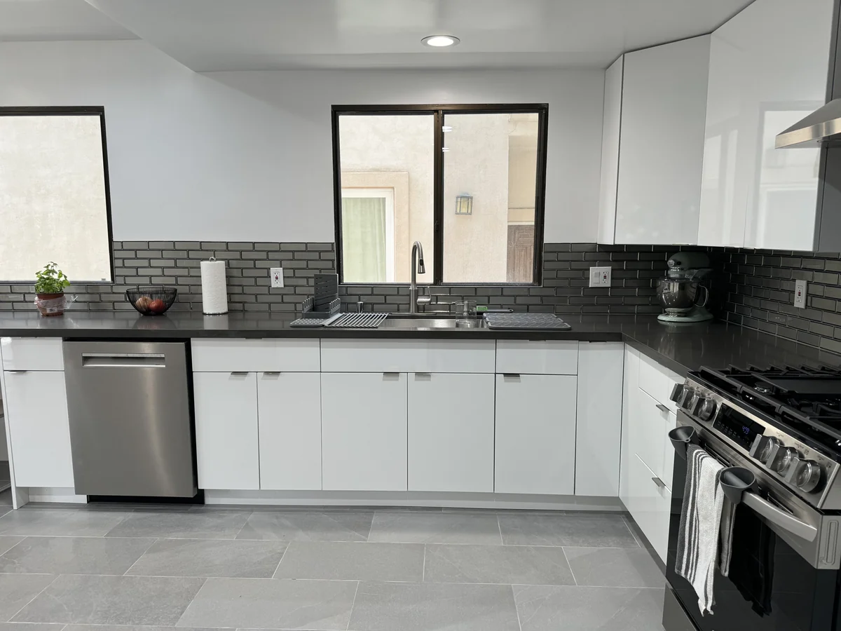 Sink beneath a window framed by glossy white cabinets and a gray glass backsplash