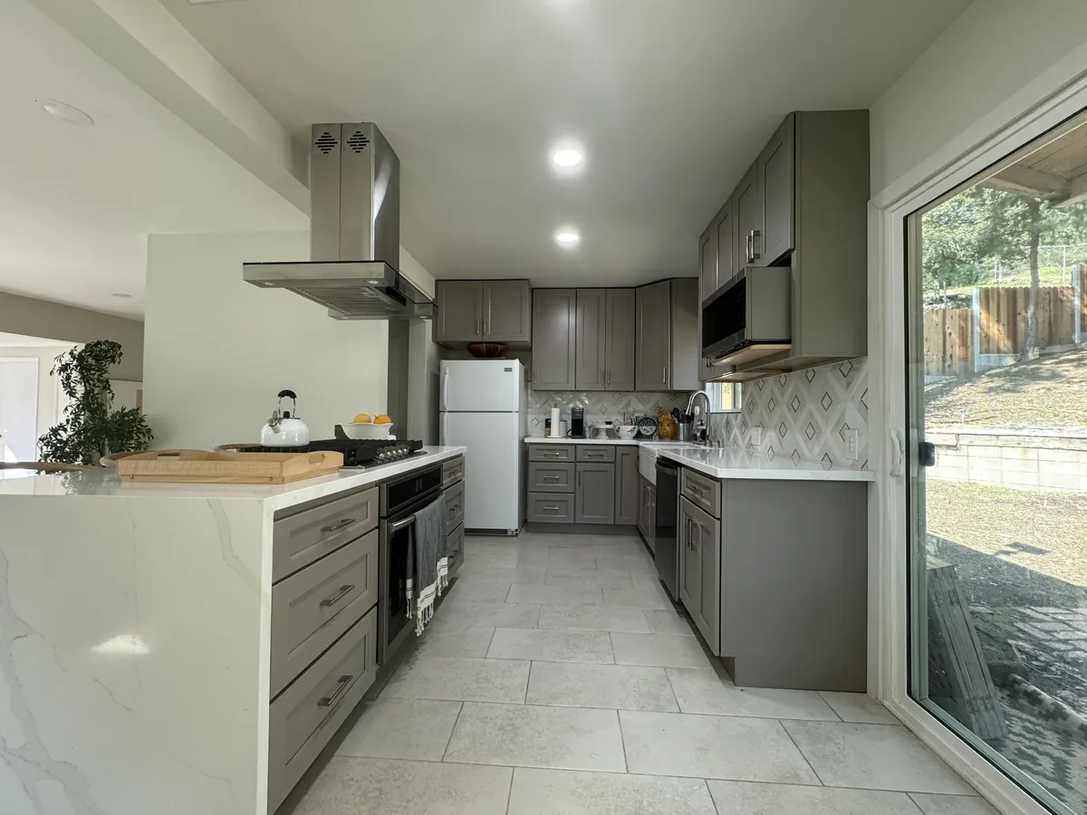 View along gray shaker cabinets toward the refrigerator with a patterned backsplash and stainless steel hood