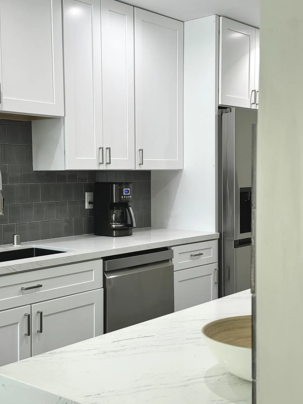 White shaker cabinets above the sink counter with the refrigerator at the end of the kitchen